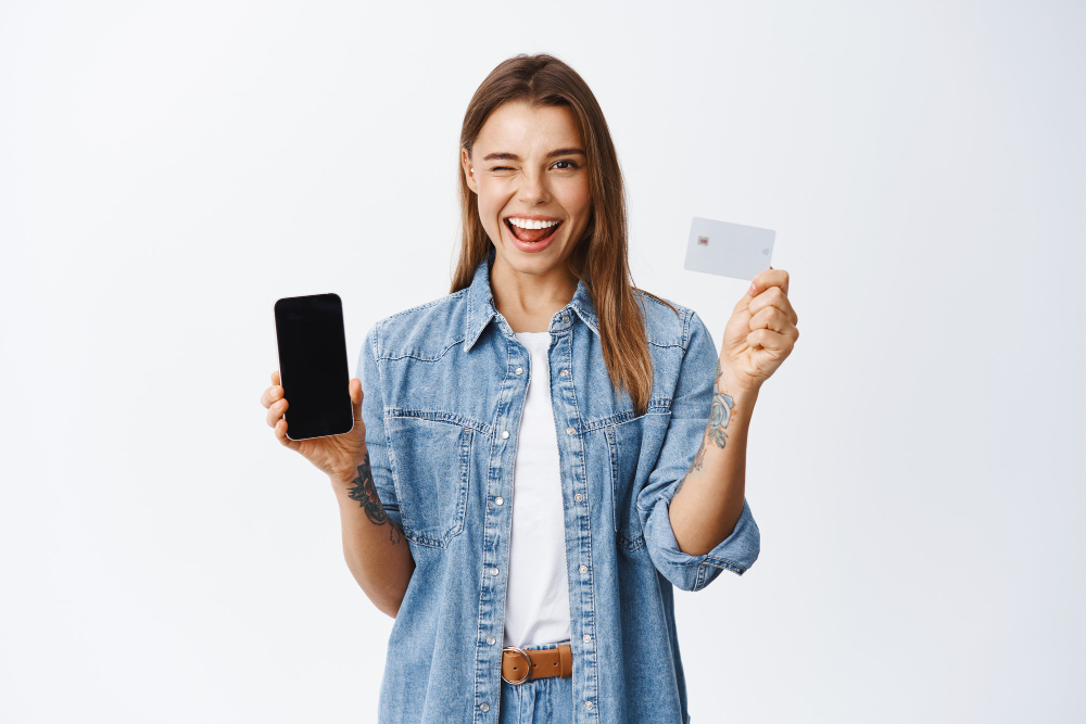 cheeky young woman winking you recommending mobile banking app showing empty smartphone screen plastic credit card white wall